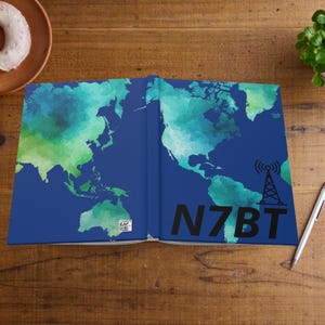 May include: A blue notebook with a world map design in shades of green and blue. The text "N7BT" and a radio tower graphic are on the cover. The notebook is open on a wooden surface.