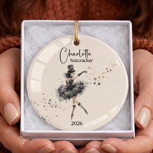 May include: A round white ceramic ornament featuring a black ink-style illustration of a ballerina in a tutu and top hat. The text reads Charlotte Nutcracker and 2026. A trail of small multi-colored stars extends from the dancer's hand.