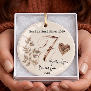 May include: A round white ceramic ornament featuring a gold string, printed with the text Hand in Hand Since 2019, a large cursive letter F, a small rope heart, a botanical branch illustration, Beautiful Years, Ken and Lori, and 2026.