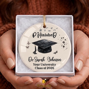 May include: A graduation ornament in a white box. The round ornament has a graduation cap graphic and the text "P Finished", "Dr. Sarah Johnson", and "Your University Class of 2026". The ornament is cream-colored with butterflies.
