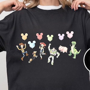 K&ouml;nnte beinhalten: Schwarzes T-Shirt mit einem farbenfrohen Aufdruck von Figuren aus einem beliebten Zeichentrickfilm. Das Design umfasst Cartoonfiguren und Mickey Mouse-f&ouml;rmige Ballons. Das Shirt hat einen Rundhalsausschnitt und kurze &Auml;rmel.