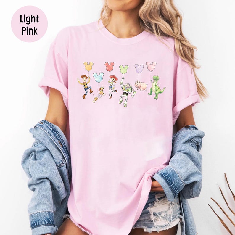 K&ouml;nnte beinhalten: Hellrosa T-Shirt mit einer Aquarellillustration von Figuren aus einem beliebten Zeichentrickfilm. Die Figuren halten Luftballons in Form von Mickey Mouse. Die Worte "Light Pink" befinden sich in einem rosa Kreis oben links.