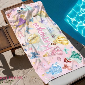 May include: A pink beach towel featuring illustrations of Disney princesses, bows, and the name "ATHRON" in a script font. The towel is draped over a wooden chaise lounge next to a pool.