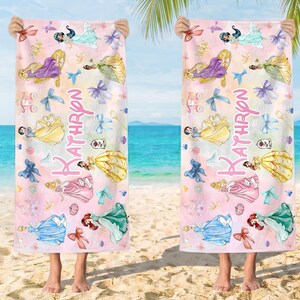 May include: Two beach towels with a pink and white watercolor design featuring various princess illustrations and the name "Kathryn" in pink script. The towels are held up by a person on a sandy beach with a view of the ocean.