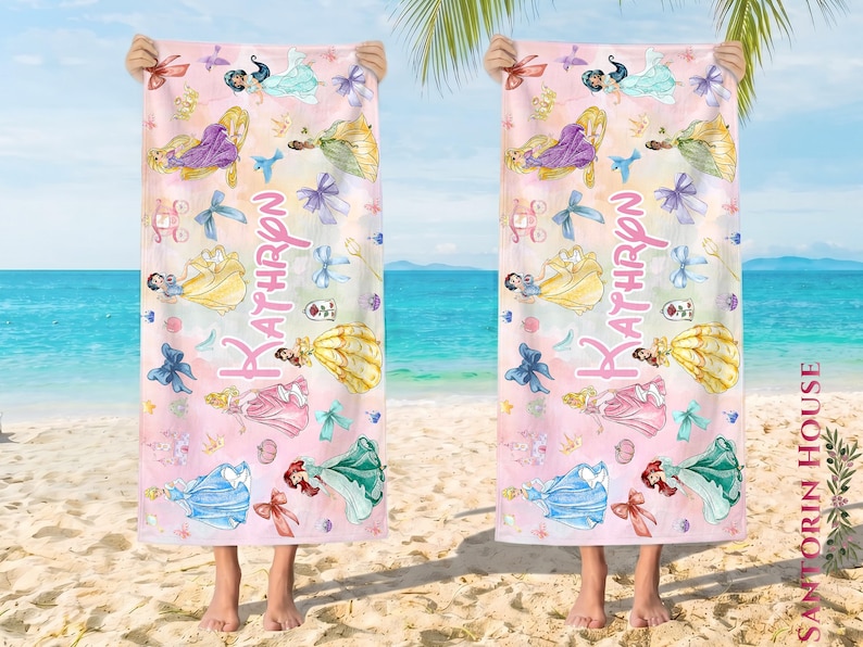 May include: Two beach towels with a pink and white watercolor design featuring various princess illustrations and the name "Kathryn" in pink script. The towels are held up by a person on a sandy beach with a view of the ocean.