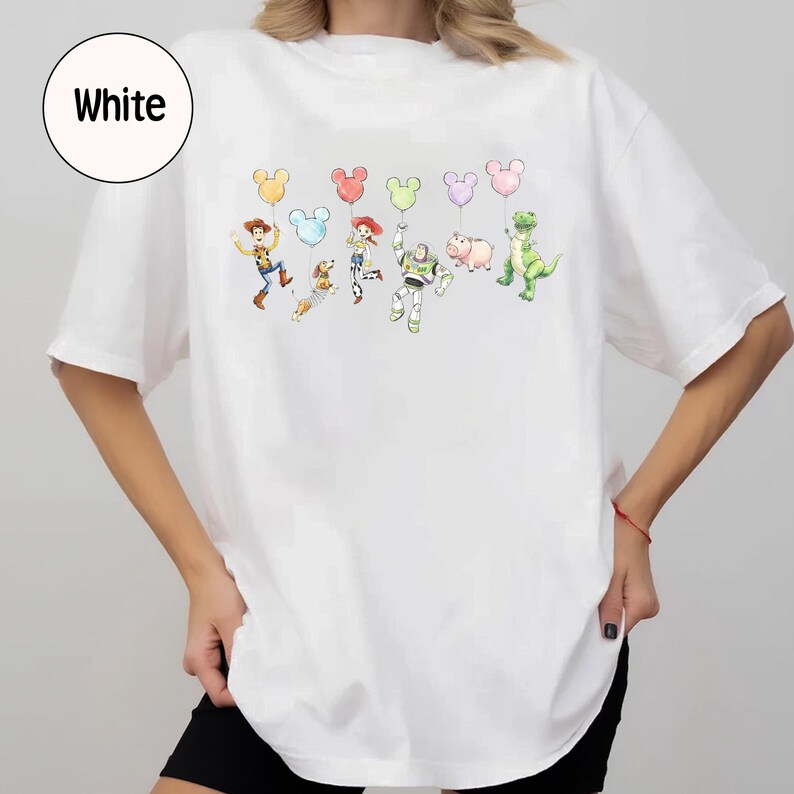 K&ouml;nnte beinhalten: Wei&szlig;es T-Shirt mit einer farbenfrohen Grafik von Zeichentrickfiguren, die Mickey Mouse-f&ouml;rmige Ballons halten. Zu den Figuren geh&ouml;ren ein Cowboy, ein Hund, ein Cowgirl, ein Astronaut, ein Schwein und ein Dinosaurier. Das Wort "White" befindet sich in einem wei&szlig;en Kreis oben links.