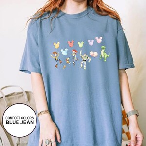 K&ouml;nnte beinhalten: Blaues Comfort Colors T-Shirt in Jeansoptik mit einer farbenfrohen Grafik von Toy Story-Charakteren und Mickey Mouse-Ballons. Das kurz&auml;rmelige Shirt hat eine lockere Passform. Der Text "COMFORT COLORS BLUE JEAN" befindet sich in einem wei&szlig;en Kreis.