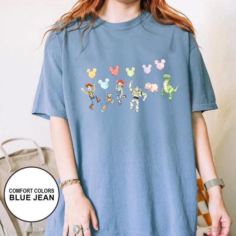 K&ouml;nnte beinhalten: Blaues Comfort Colors T-Shirt in Jeansoptik mit einer farbenfrohen Grafik von Toy Story-Charakteren und Mickey Mouse-Ballons. Das kurz&auml;rmelige Shirt hat eine lockere Passform. Der Text "COMFORT COLORS BLUE JEAN" befindet sich in einem wei&szlig;en Kreis.