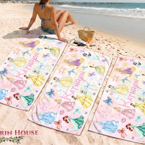 May include: Three pink beach towels with princess illustrations and the name "KATHRYN" in pink script. The towels feature images of princesses in various gowns, bows, and other decorative elements. The towels are on a sandy beach.