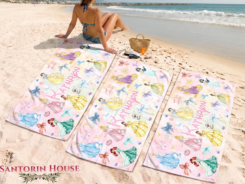 May include: Three pink beach towels with princess illustrations and the name "KATHRYN" in pink script. The towels feature images of princesses in various gowns, bows, and other decorative elements. The towels are on a sandy beach.