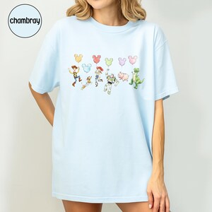 K&ouml;nnte beinhalten: Hellblaues T-Shirt mit einer Cartoon-Grafik von Figuren und Mickey Mouse-f&ouml;rmigen Luftballons. Zu den Figuren geh&ouml;ren ein Cowboy, ein Weltraumranger, ein Cowgirl, ein Schwein, ein Dinosaurier und ein Hund. Das Wort "chambray" befindet sich in einem Kreis oben links.