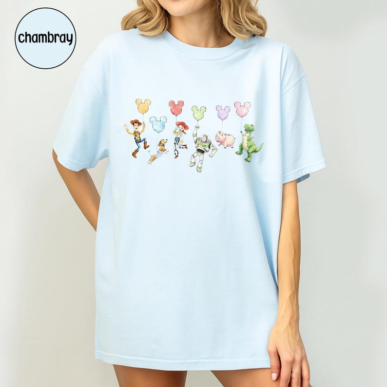 K&ouml;nnte beinhalten: Hellblaues T-Shirt mit einer Cartoon-Grafik von Figuren und Mickey Mouse-f&ouml;rmigen Luftballons. Zu den Figuren geh&ouml;ren ein Cowboy, ein Weltraumranger, ein Cowgirl, ein Schwein, ein Dinosaurier und ein Hund. Das Wort "chambray" befindet sich in einem Kreis oben links.