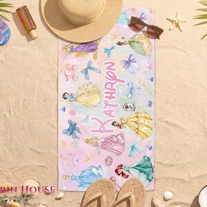 May include: A rectangular beach towel with a watercolor design featuring Disney princesses in various gowns. The towel has a pink and white ombre background with the name "Kathryn" in pink script. Other beach accessories are in the background.