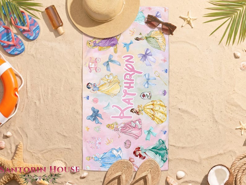 May include: A rectangular beach towel with a watercolor design featuring Disney princesses in various gowns. The towel has a pink and white ombre background with the name "Kathryn" in pink script. Other beach accessories are in the background.