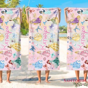 May include: Three beach towels with a pink and white watercolor design featuring princess illustrations, bows, and the name "Kathryn". The towels are held up against a beach backdrop.