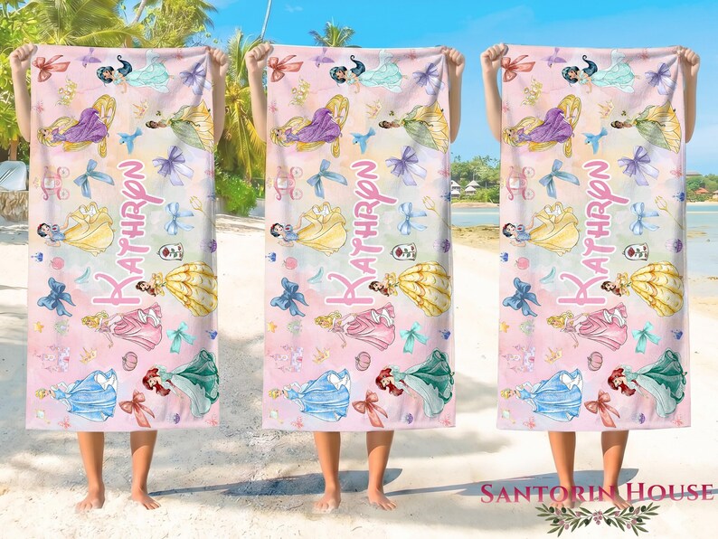 May include: Three beach towels with a pink and white watercolor design featuring princess illustrations, bows, and the name "Kathryn". The towels are held up against a beach backdrop.