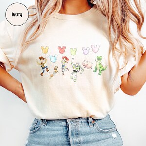 K&ouml;nnte beinhalten: Elfenbeinfarbenes T-Shirt mit einer Aquarellillustration von Toy Story-Charakteren, die Mickey Mouse-f&ouml;rmige Ballons in verschiedenen Farben halten. Das Shirt hat einen Rundhalsausschnitt und kurze &Auml;rmel. Das Wort "ivory" befindet sich in einem Kreis oben links.