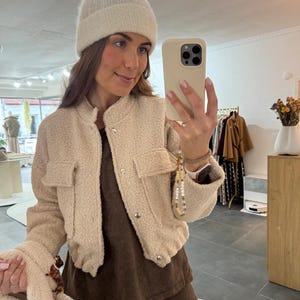 Könnte beinhalten: Beige Jacke mit Textur, zwei Vordertaschen und silbernen Knöpfen. Die Jacke wird mit einer passenden Mütze und einem braunen Oberteil kombiniert. Die Person hält ein Telefon in einer beigen Hülle.