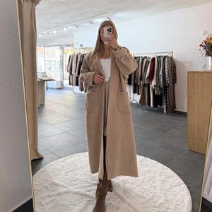 Könnte beinhalten: Ein langer, beigefarbener Mantel mit großen Vordertaschen und einer lockeren Passform. Der Mantel ist offen und zeigt ein weißes Oberteil und einen langen Rock. Die Person im Bild trägt braune Stiefel. Der Hintergrund zeigt ein Bekleidungsgeschäft mit Kleiderständern.