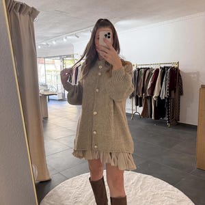 Könnte beinhalten: Ein beiges Strick-Cardigan-Kleid mit großen perlenartigen Knöpfen. Das Kleid hat einen Rüschensaum und wird mit braunen kniehohen Stiefeln kombiniert. Im Hintergrund ist eine Kleiderstange mit verschiedenen Kleidungsstücken und ein Spiegel zu sehen.