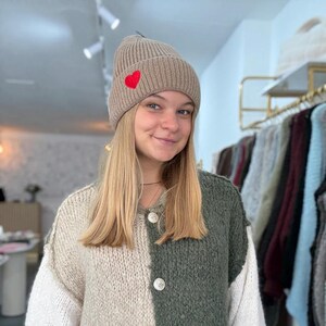 Könnte beinhalten: Eine beige Strickmütze mit einem roten Herzaufnäher. Die Mütze ist gerippt und hat einen umgeschlagenen Rand. Die Person trägt einen zweifarbigen Pullover in Creme und Olivgrün.