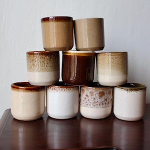 Pode incluir: Uma coleção de dez pequenas chávenas cilíndricas de cerâmica em tons variados de castanho e branco. As chávenas têm um aspeto rústico e artesanal, com um acabamento vidrado e uma forma lisa e arredondada. As chávenas estão empilhadas.