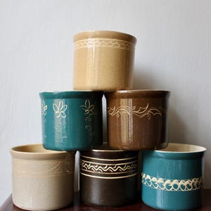 Könnte beinhalten: Ein Stapel von sechs Keramiktöpfen in verschiedenen Farben, darunter Beige, Türkis und Braun. Jeder Topf hat ein dekoratives Band mit bemalten Designs, wie Blumenmustern und wellenförmigen Linien. Die Töpfe sind zylindrisch mit einem leicht ausgestellten Rand.