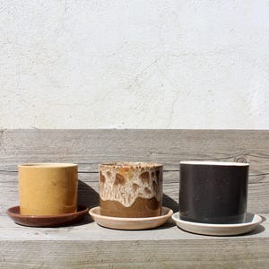 Könnte beinhalten: Drei Keramik-Blumentöpfe mit Untersetzern. Der erste ist senfgelb, der zweite braun mit weißer Glasur und der dritte dunkelbraun mit weißem Rand. Sie stehen auf einer verwitterten Holzoberfläche.