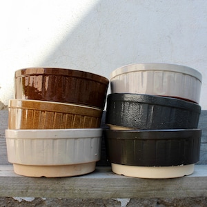 May include: A stack of six ceramic ramekins in various colors: dark brown, tan, cream, and black. Each ramekin has a ribbed exterior and a rounded shape, perfect for baking and serving individual portions.