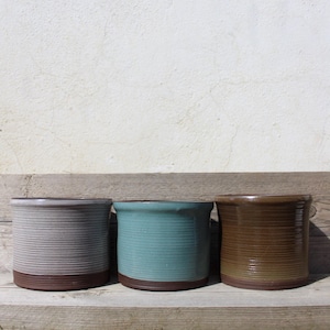 Op de afbeelding: Drie keramische bloempotten op een rij, elk met een andere kleur en textuur. De eerste is grijs met horizontale ribbels en een bruine basis. De tweede is blauwgroen met verticale ribbels en een bruine basis. De derde is bruin met verticale ribbels.