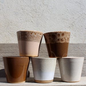 May include: A collection of five ceramic cups in various earth-tone colors. The cups have a tapered design and a speckled or glazed finish. Colors include light pink, brown, white, and beige. The cups are arranged on a wooden surface.