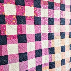 May include: A quilt with a checkered pattern in shades of pink, white, and navy blue. The quilt is quilted with a decorative design of curved lines.