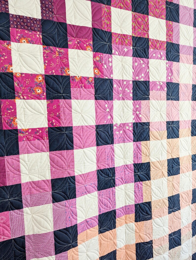 May include: A quilt with a checkered pattern in shades of pink, white, and navy blue. The quilt is quilted with a decorative design of curved lines.