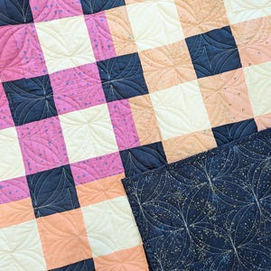 May include: A quilt with a checkered pattern in pink, peach, white, and navy blue. The quilt is quilted with a swirling pattern.
