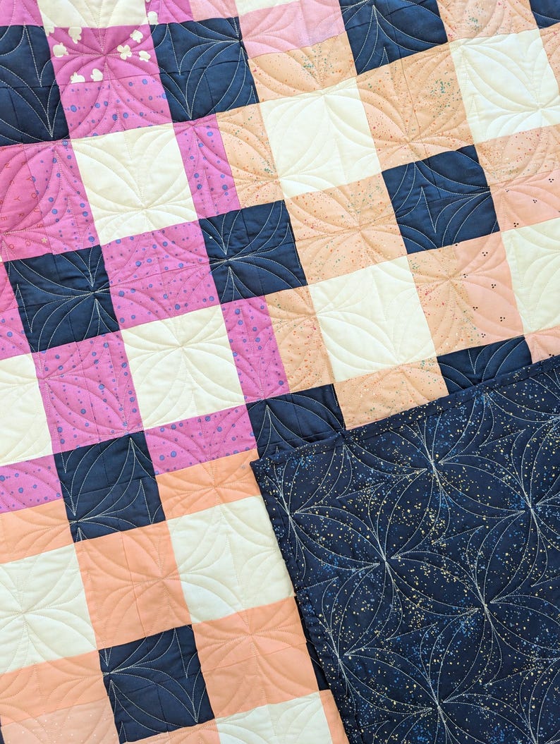 May include: A quilt with a checkered pattern in pink, peach, white, and navy blue. The quilt is quilted with a swirling pattern.