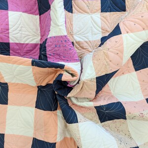 May include: A quilt with a checkered pattern in pink, peach, white, and navy blue. The quilt is quilted with a swirling leaf design.