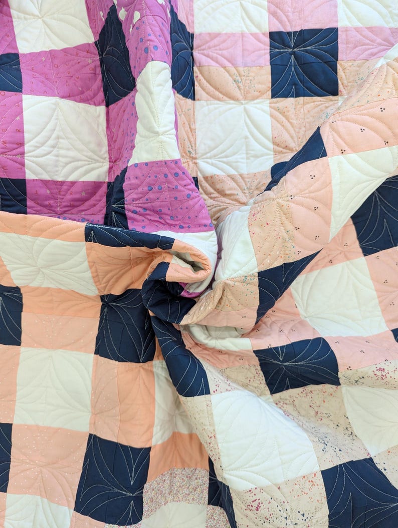 May include: A quilt with a checkered pattern in pink, peach, white, and navy blue. The quilt is quilted with a swirling leaf design.