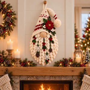 May include: A cream-coloured crocheted gnome wall hanging featuring a tall pointed hat with red stripes and a pom-pom. The gnome has a textured beard adorned with red berries, green leaves, a red poinsettia, white snowflakes, and small pinecones.