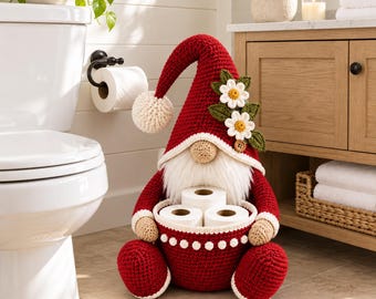 Pode incluir: Uma figura de gnomo em crochê nas cores vermelho e bege, com um chapéu alto e pontudo adornado com flores brancas e folhas verdes. O gnomo tem uma longa barba branca e segura uma cesta redonda contendo três rolos de papel higiênico.