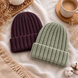 Może przedstawiać: Dwie czapki typu beanie z dzianiny o prążkowanej fakturze z wywiniętym mankietem. Jedna czapka jest w kolorze głębokiego śliwkowego fioletu, a druga w kolorze delikatnej szałwiowej zieleni. Obie czapki mają gruby splot z wyraźnymi pionowymi liniami.