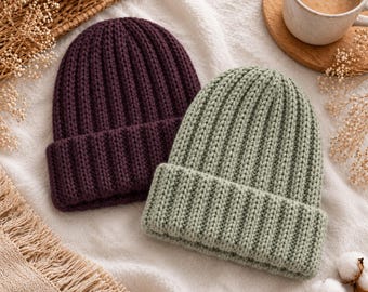 Pode incluir: Dois gorros de malha com textura canelada e barra dobrada. Um gorro é de cor roxo ameixa profundo e o outro é de cor verde sálvia suave. Ambos os gorros são construídos com um padrão de malha de fio grosso, exibindo linhas verticais.