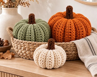 Pode incluir: Três abóboras de crochê nas cores verde sálvia, laranja brilhante e creme, cada uma com um caule marrom escuro. As abóboras estão dispostas em uma cesta de vime trançada, com as abóboras verde e laranja dentro e a creme colocada à frente.
