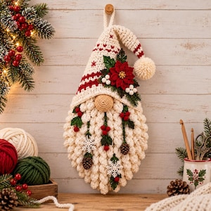 May include: A crocheted Christmas gnome wall hanging featuring a cream-coloured hat with red stripes, a large cream beard, a central tan nose, and decorative accents including a red poinsettia, green leaves, white berries, pinecones, and snowflakes.