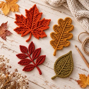 May include: Four crocheted leaves in orange, mustard yellow, dark red, and olive green. The leaves feature visible yarn textures and central veins. A wooden crochet hook and a length of twine are positioned near the handmade leaf appliques.