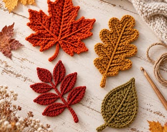 May include: Four crocheted leaves in orange, mustard yellow, dark red, and olive green. The leaves feature visible yarn textures and central veins. A wooden crochet hook and a length of twine are positioned near the handmade leaf appliques.