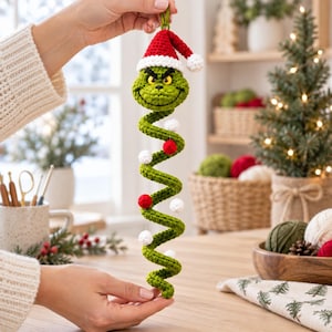 Könnte beinhalten: Ein gehäkeltes grünes Spiralornament mit einem Charakterkopf mit gelben Augen und einer rot-weißen Weihnachtsmannmütze. Der lange, gewundene Körper ist mit kleinen, abwechselnd roten und weißen Pompons verziert, die entlang der Spirale befestigt sind.