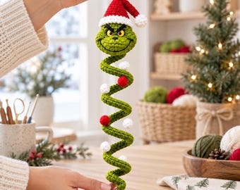 Pode incluir: Um ornamento em espiral verde de crochê com uma cabeça de personagem de olhos amarelos e um gorro de Papai Noel vermelho e branco. O corpo longo e enrolado é decorado com pequenos pompons vermelhos e brancos alternados ao longo da espiral.