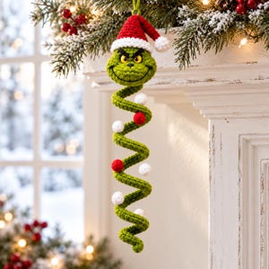 Könnte beinhalten: Ein grünes, gehäkeltes Spiral-Hängeornament mit einem Charakterkopf, der eine rot-weiße Weihnachtsmannmütze trägt. Der lange, gewundene Körper ist mit kleinen, abwechselnd roten und weißen gehäkelten Kugeln entlang der Spirale verziert.