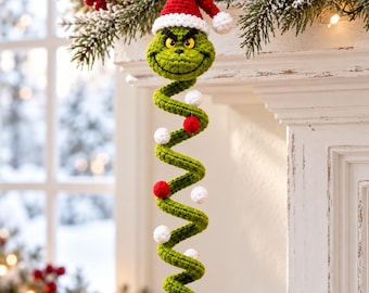 Pode incluir: Um ornamento suspenso em espiral de crochê verde com uma cabeça de personagem usando um gorro de Papai Noel vermelho e branco. O corpo longo e enrolado é decorado com pequenas esferas de crochê vermelhas e brancas alternadas.