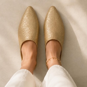 Puede incluir: Un par de zapatos sin cordones de color dorado con punta afilada, que presentan un sutil y complejo patrón floral en relieve en toda la superficie superior. Los zapatos tienen un diseño de perfil bajo con punta cerrada y talón abierto.